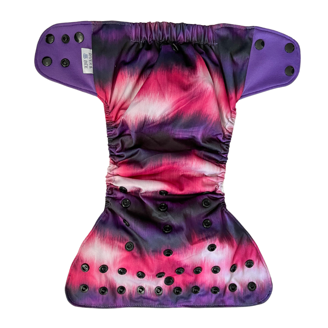 Photo: River & Bee modern cloth nappy in the print Aura. Flatlay showing the outside of the nappy. Pink and purple with black snaps.